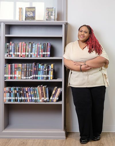 Woman with blonde updo wearing red glasses smiles, gray shirt with blue flowers; books in background.