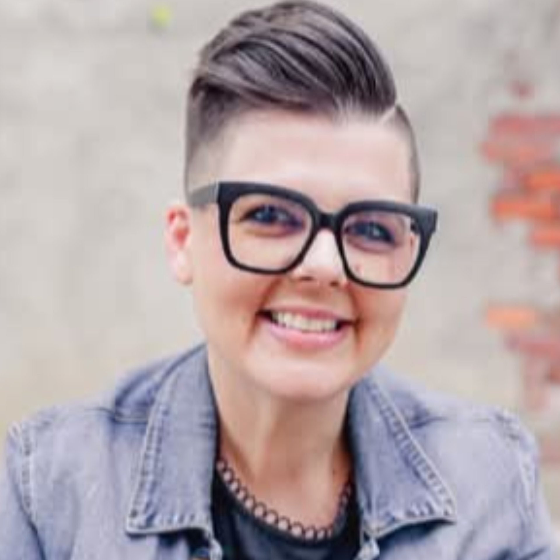 Woman with short hair and large glasses smiles, wearing denim jacket and a necklace.