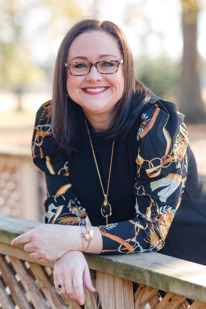 Woman with glasses smiles while leaning on a wooden fence, wearing a black dress with patterned sleeves.