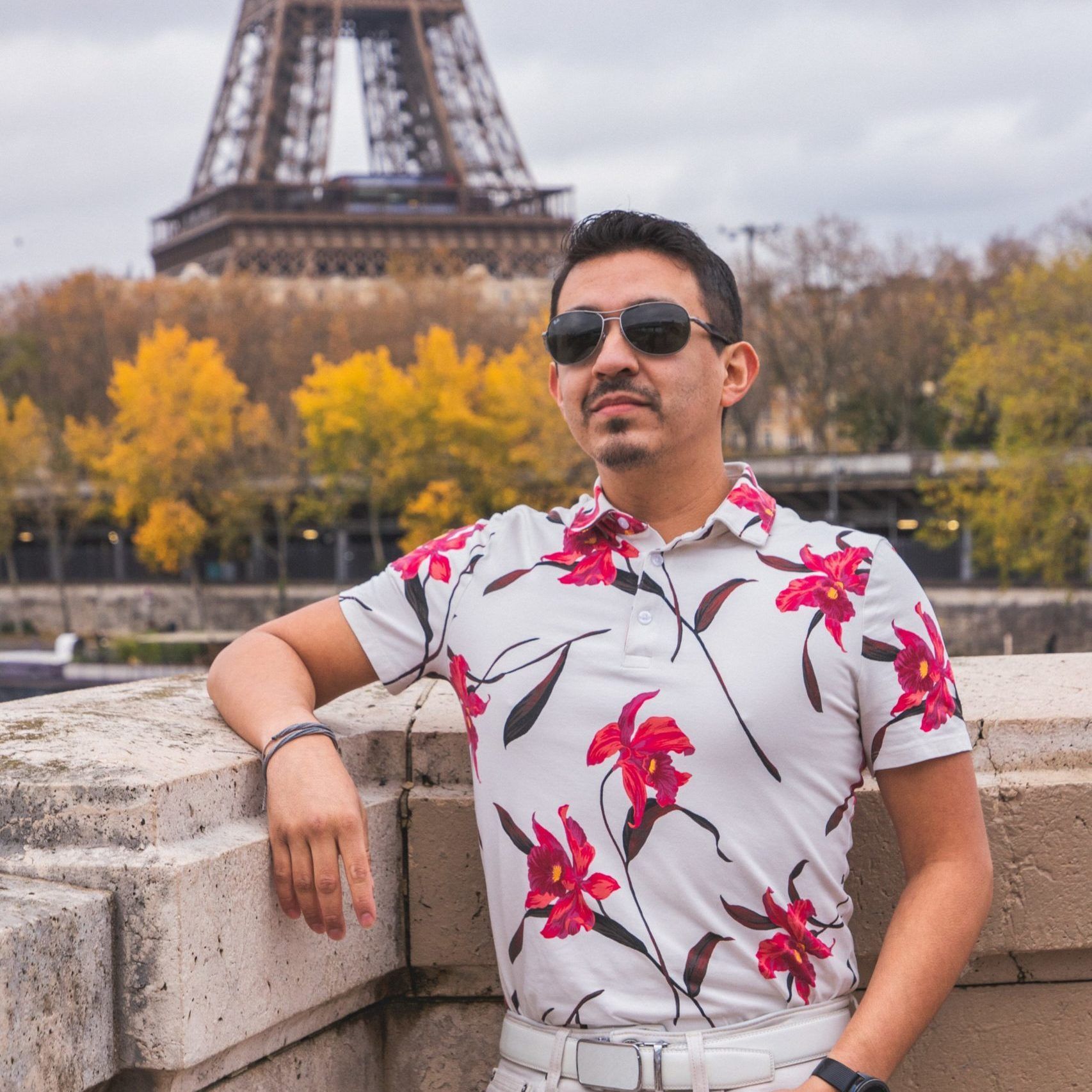 Man in floral shirt, sunglasses, leaning on a stone ledge in front of the Eiffel Tower, Paris.
