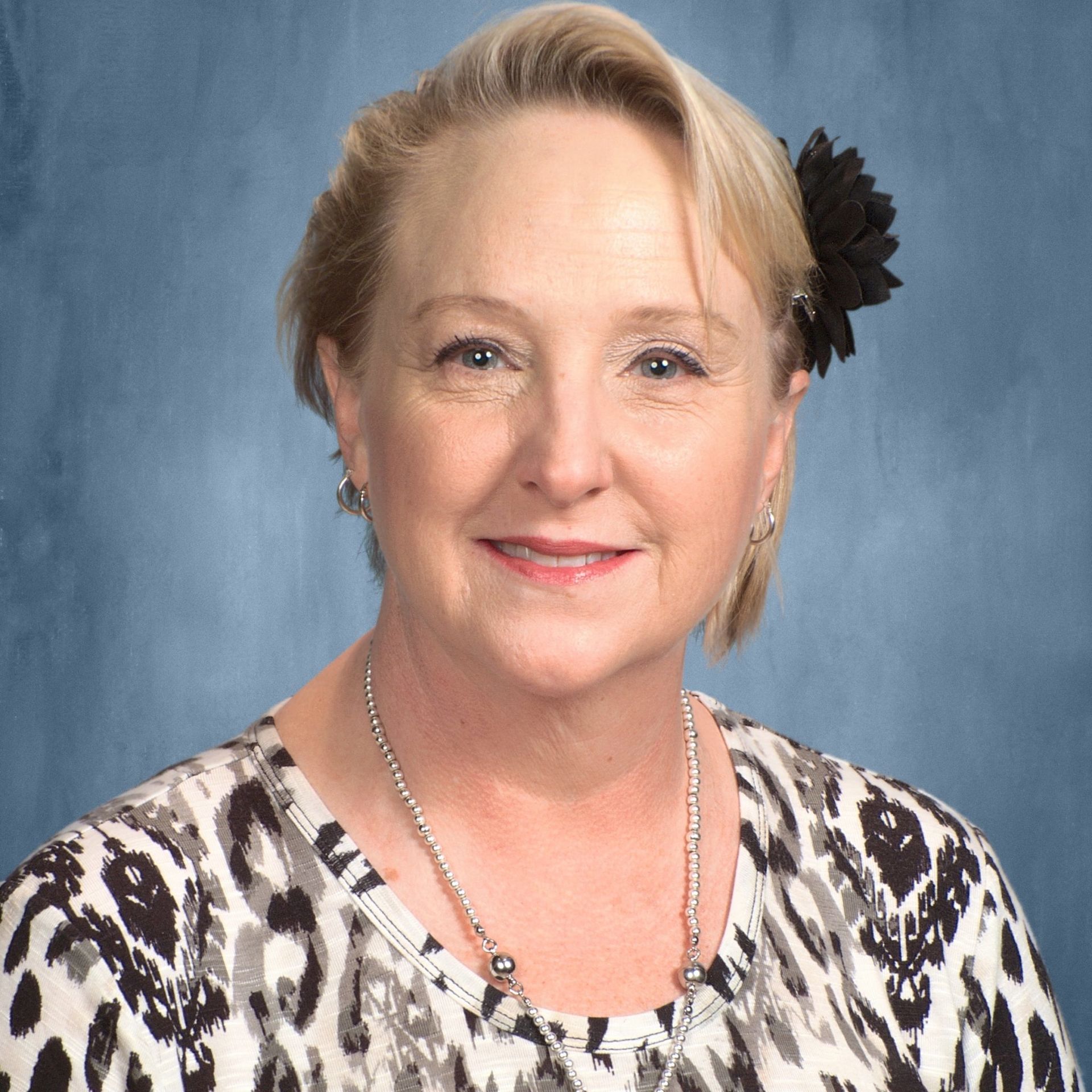 Woman with blonde hair wearing a patterned shirt and a black flower hair accessory, smiling.