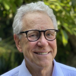 Man with gray hair and glasses smiles outdoors.