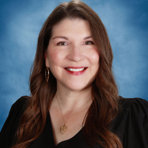 Woman with long brown hair, smiling, wearing a black top and gold jewelry, against a blue background.