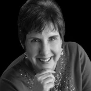 Woman smiling, resting chin on hand, wearing a dark top with embellishments, against a black background.