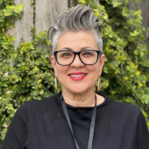 Woman with gray hair, glasses, and red lipstick smiling, wearing black, with greenery background.