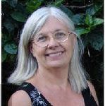Woman with gray hair and glasses smiling in front of green foliage.