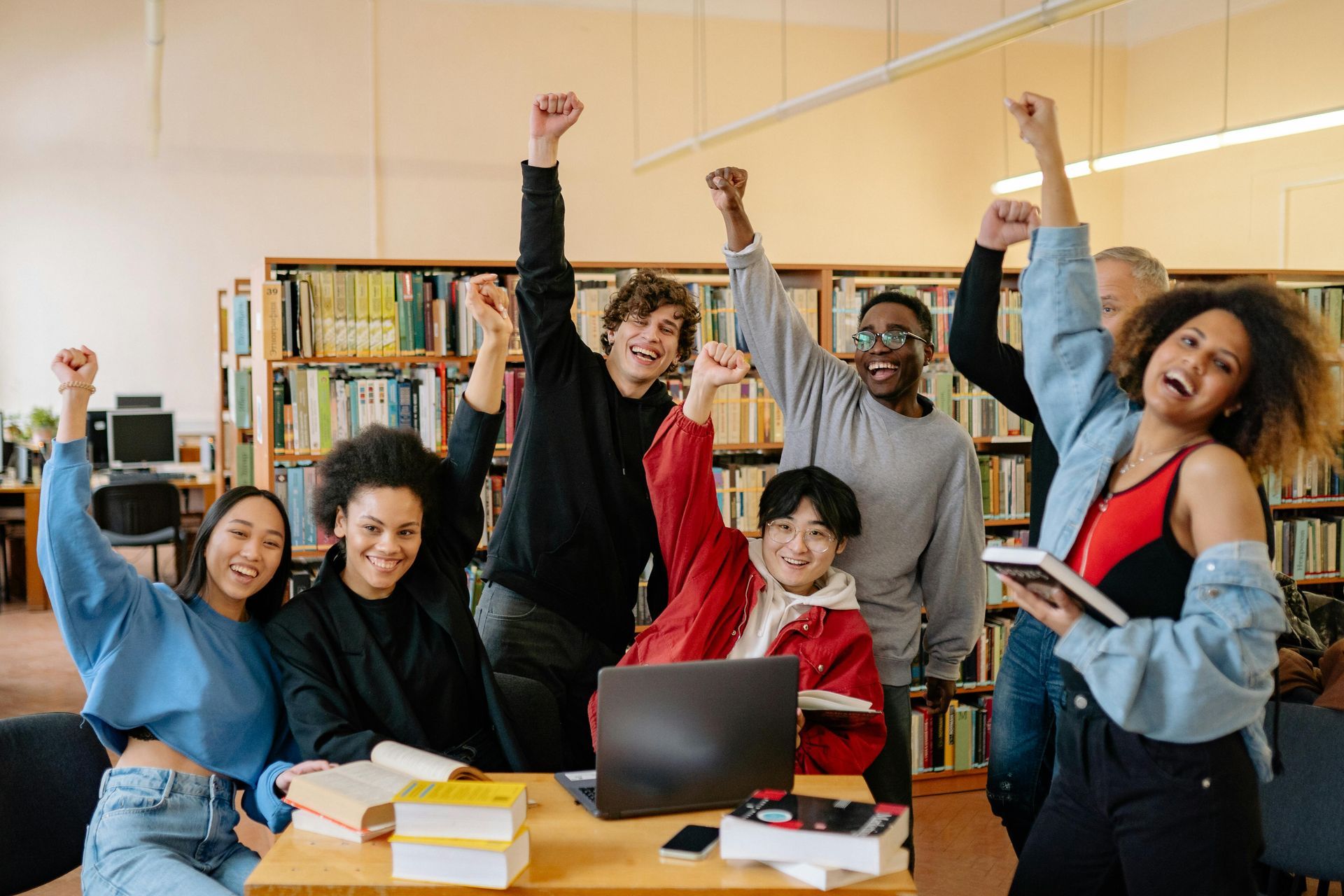 Group of people celebrating with raised fists in a library, smiling and holding books.