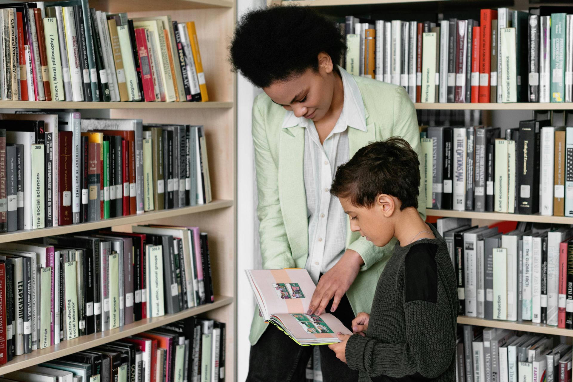 Woman and child looking at a book in a library, surrounded by bookshelves.