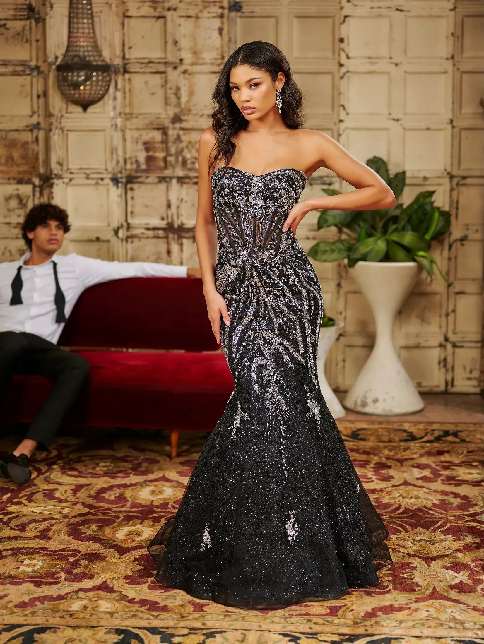 Woman in strapless sequin gown, posing in front of a red couch. Man in a suit sits on the couch.