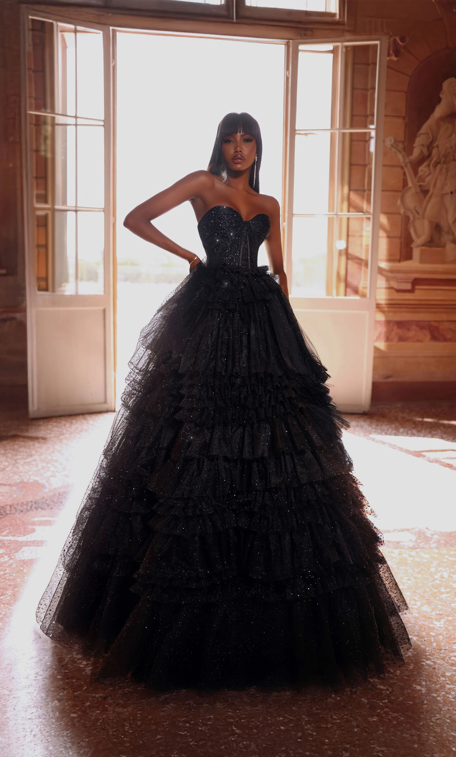 Woman in strapless black gown with a layered, full skirt; standing in front of a window.