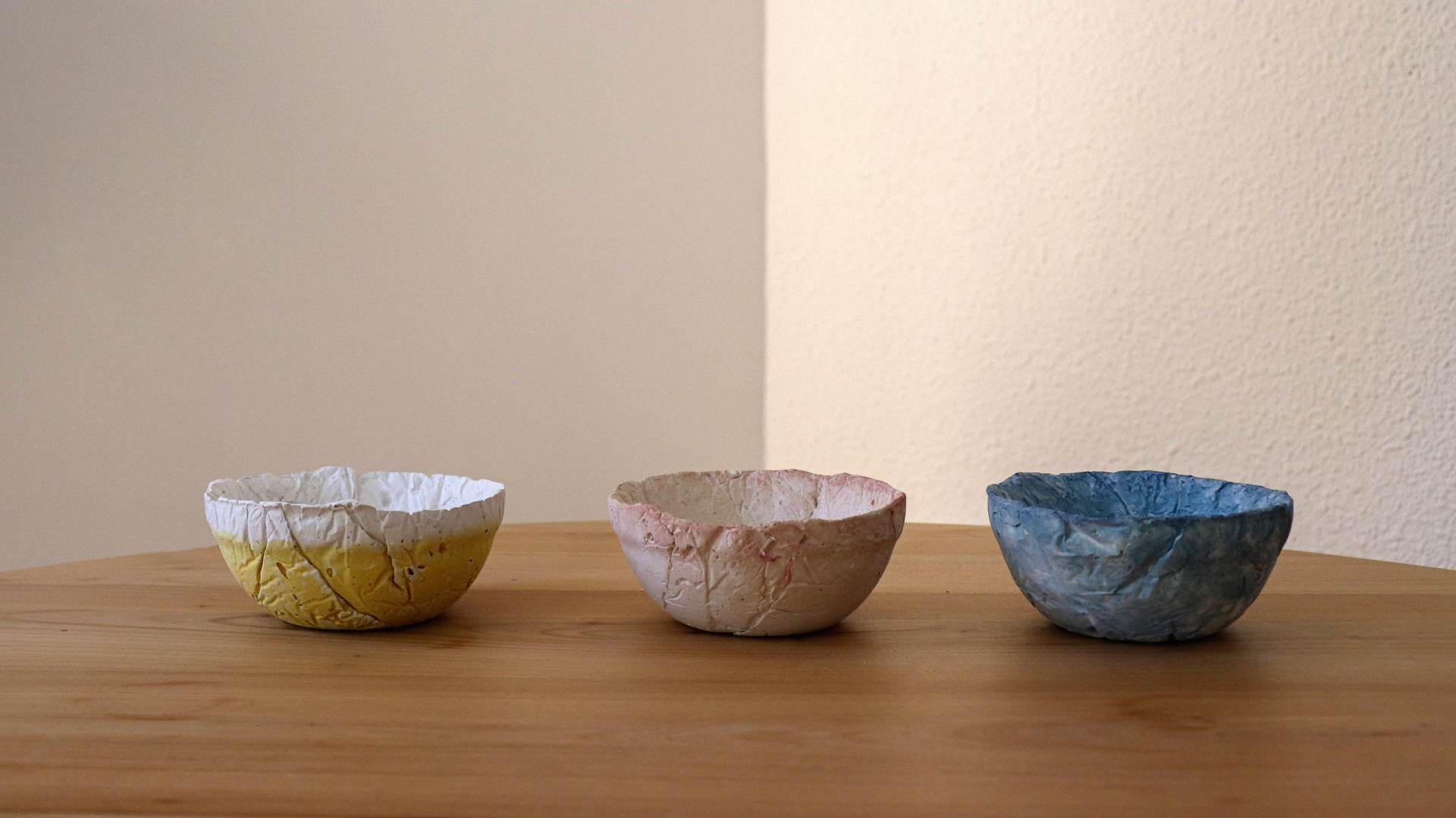 Fotografia dels productes final del projecte egg dishes fets amb biomaterial. Recolzats sobre una taula de fusta i fons blanc. Dissenyat per mersi studio. Vídeo agafant ous i preparant-los per a la seva posterior producció a la cuina d'una casa, ja que després seran ficats al forn. Dissenyat per mersi studio.
