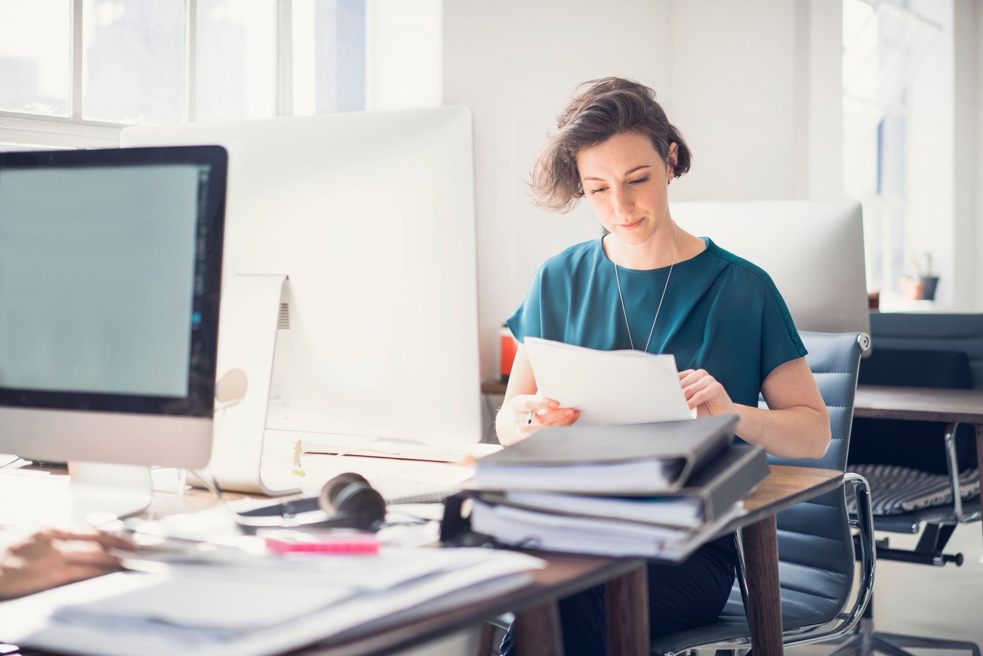 A woman is sitting at a desk in front of a computer holding a piece of paper.