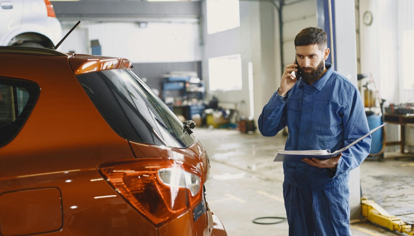 Mechanic in a blue uniform talks on phone beside a red car in an auto repair shop, highlighting the realities of franchise ownership.