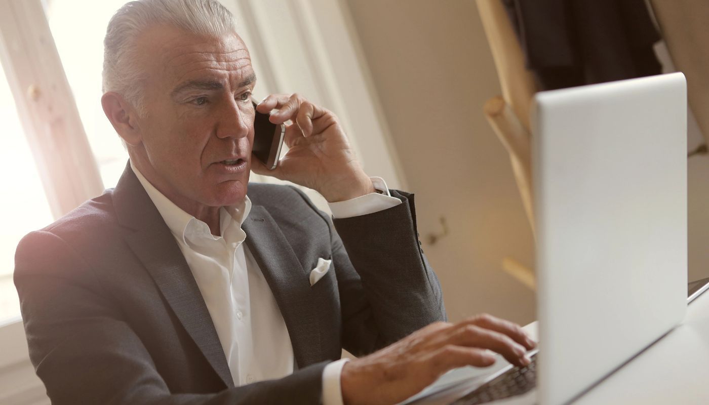 Senior businessman managing a phone call while working on a laptop in a modern office, illustrating semi-absentee franchise ownership