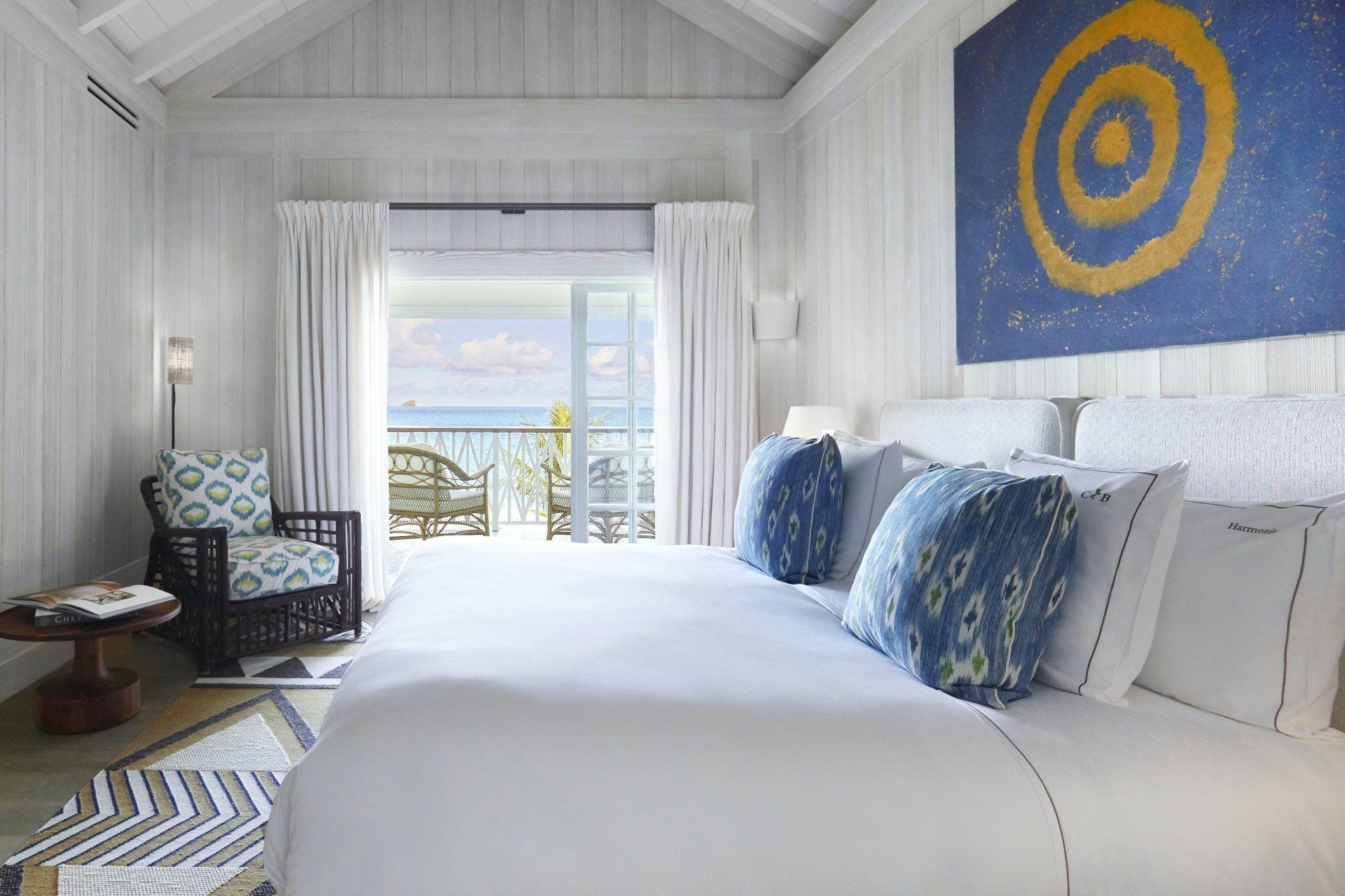 Bedroom with white walls, bed, and artwork; opens to a balcony overlooking the ocean.