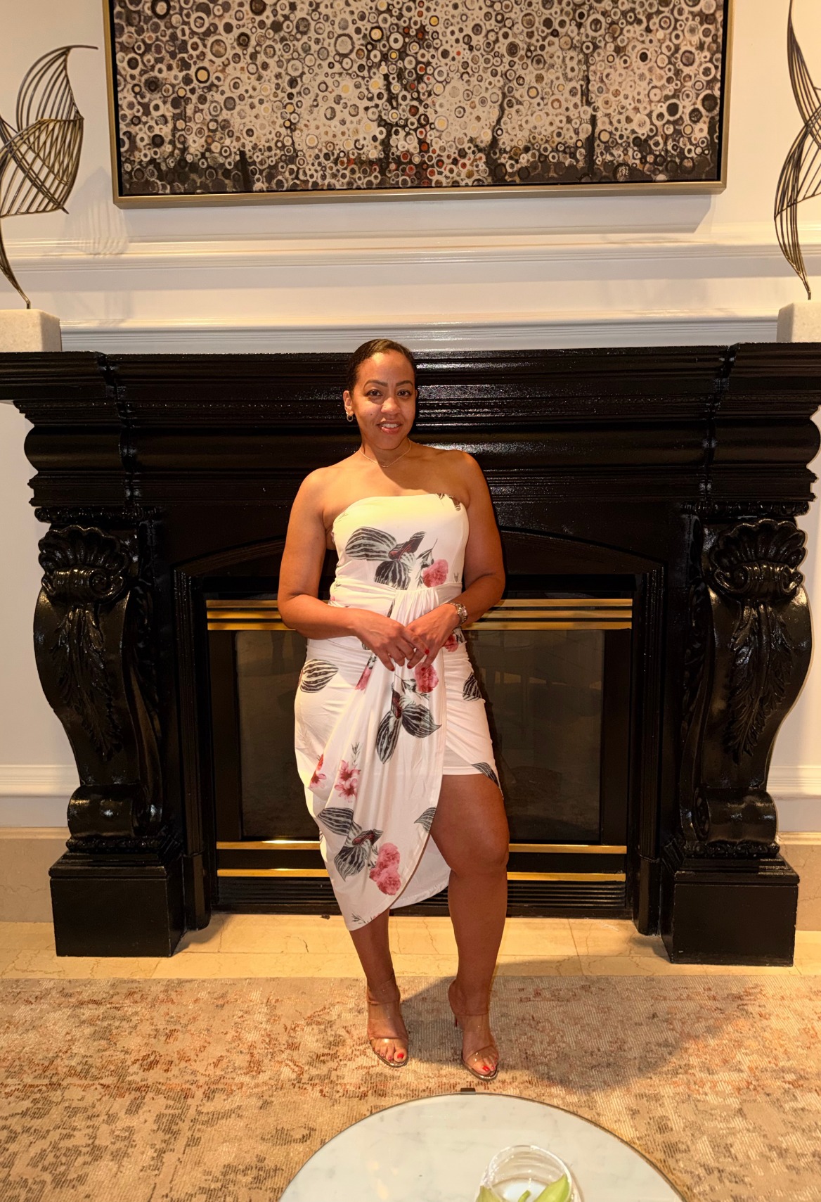 Woman posing in a white floral dress in front of a black fireplace and ornate wall art.