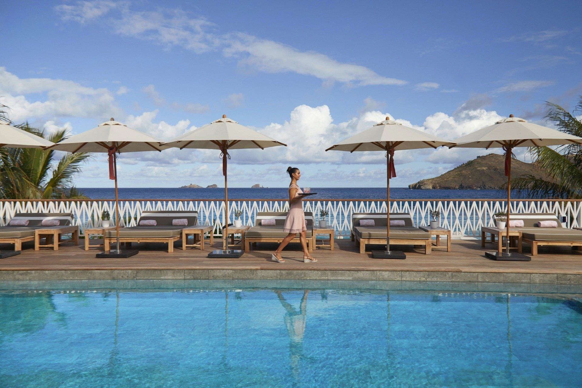 Poolside scene: Person walks past lounge chairs, umbrellas, pool, and ocean view.