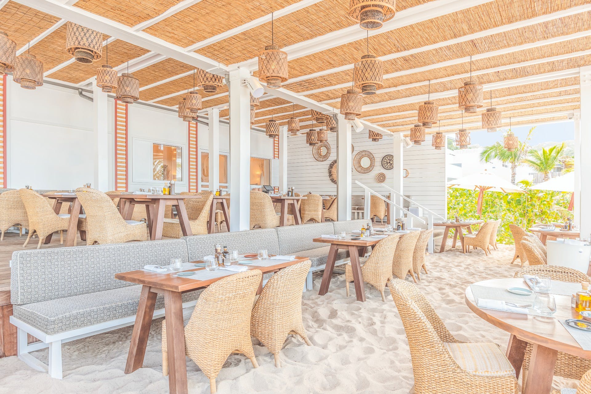 Beachside restaurant with woven chairs, tables, and a sand floor under a straw roof.