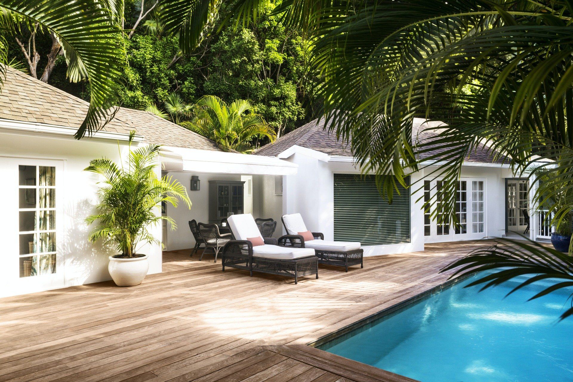 White house with pool, wooden deck, lounge chairs. Lush green foliage.