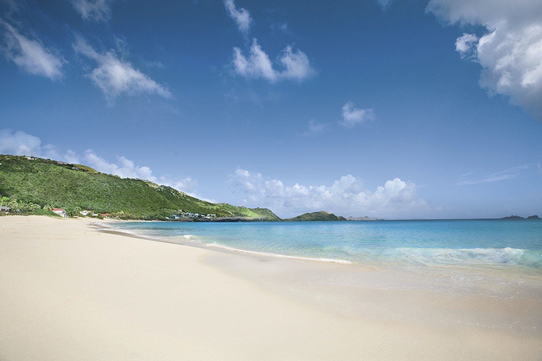White sand beach with turquoise water, lush green hillside, blue sky with white clouds.