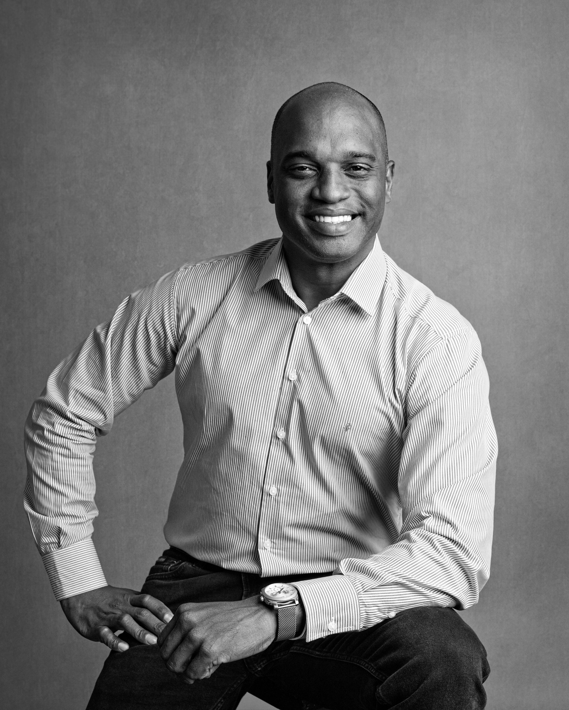 Black and white portrait of a smiling Black man in a striped shirt, seated with hands clasped.