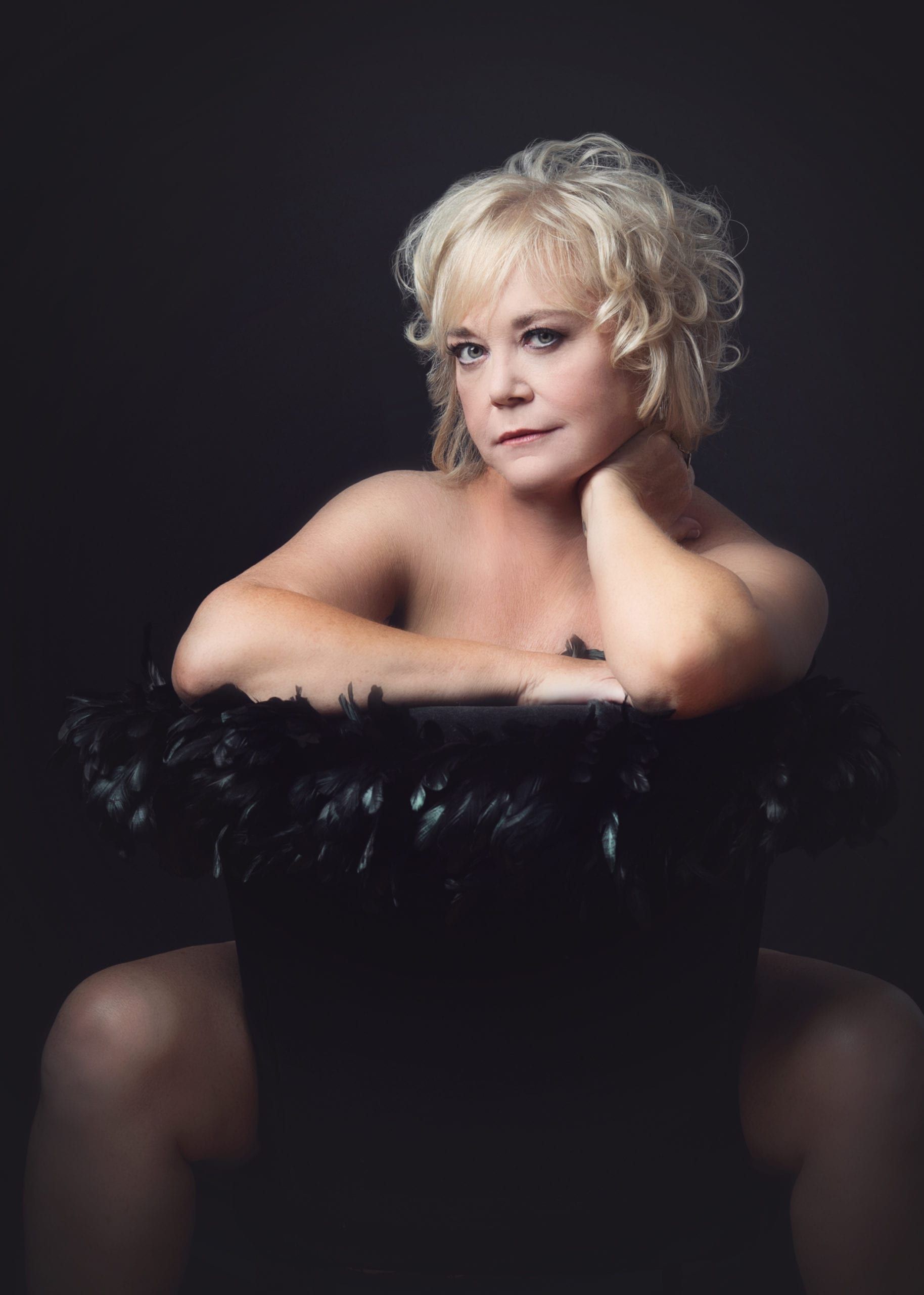Woman with blonde hair, sitting on a black chair with feather trim. She looks toward the viewer with a slight smile.