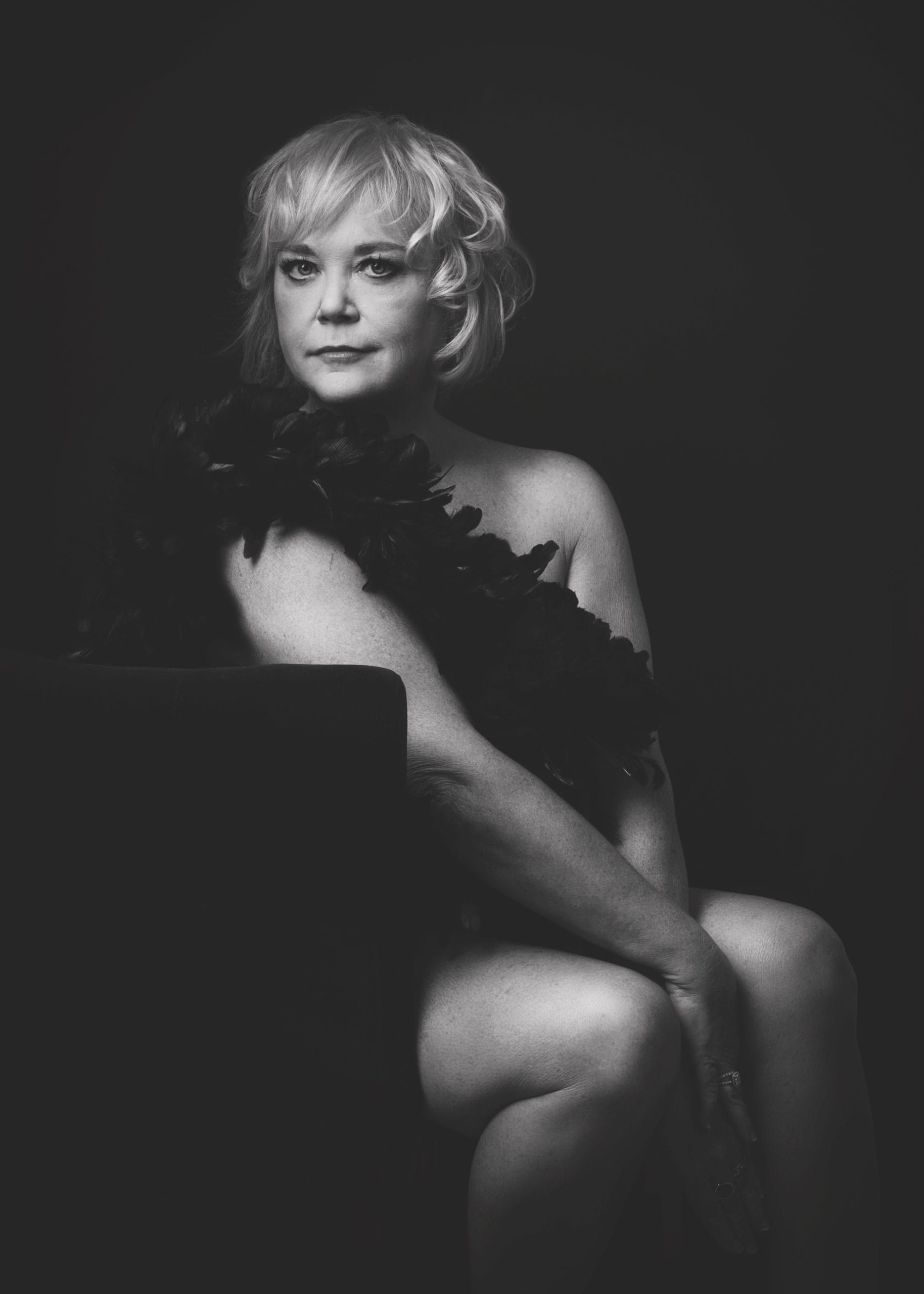 Woman with short blonde hair, draped in a black boa. Posing, in a studio. Black and white photo.