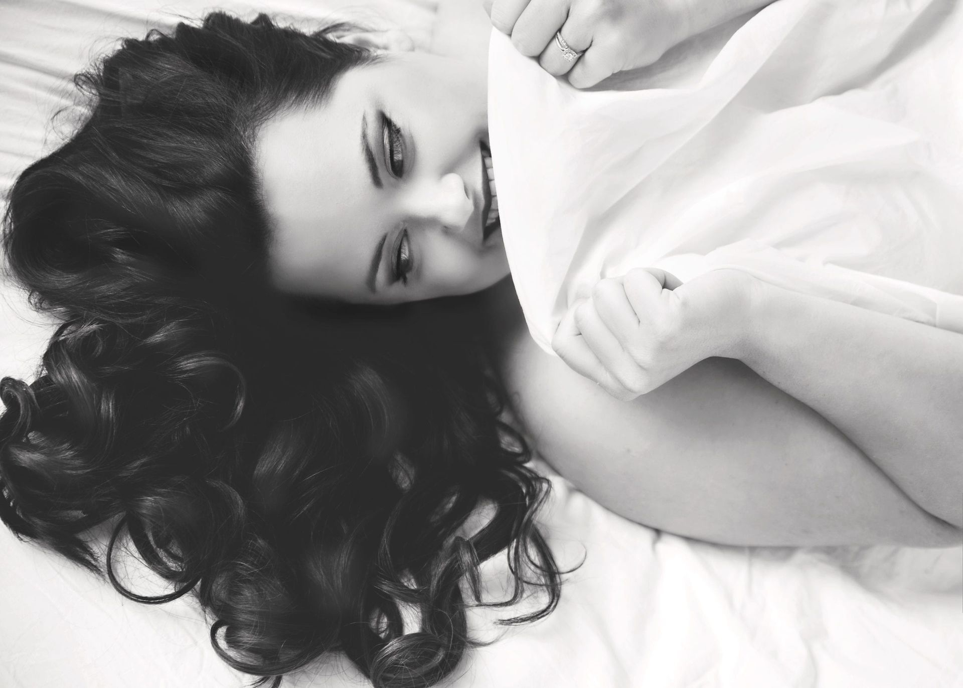 Woman with dark curly hair smiles, lying on white sheets, holding sheet close. Black and white.