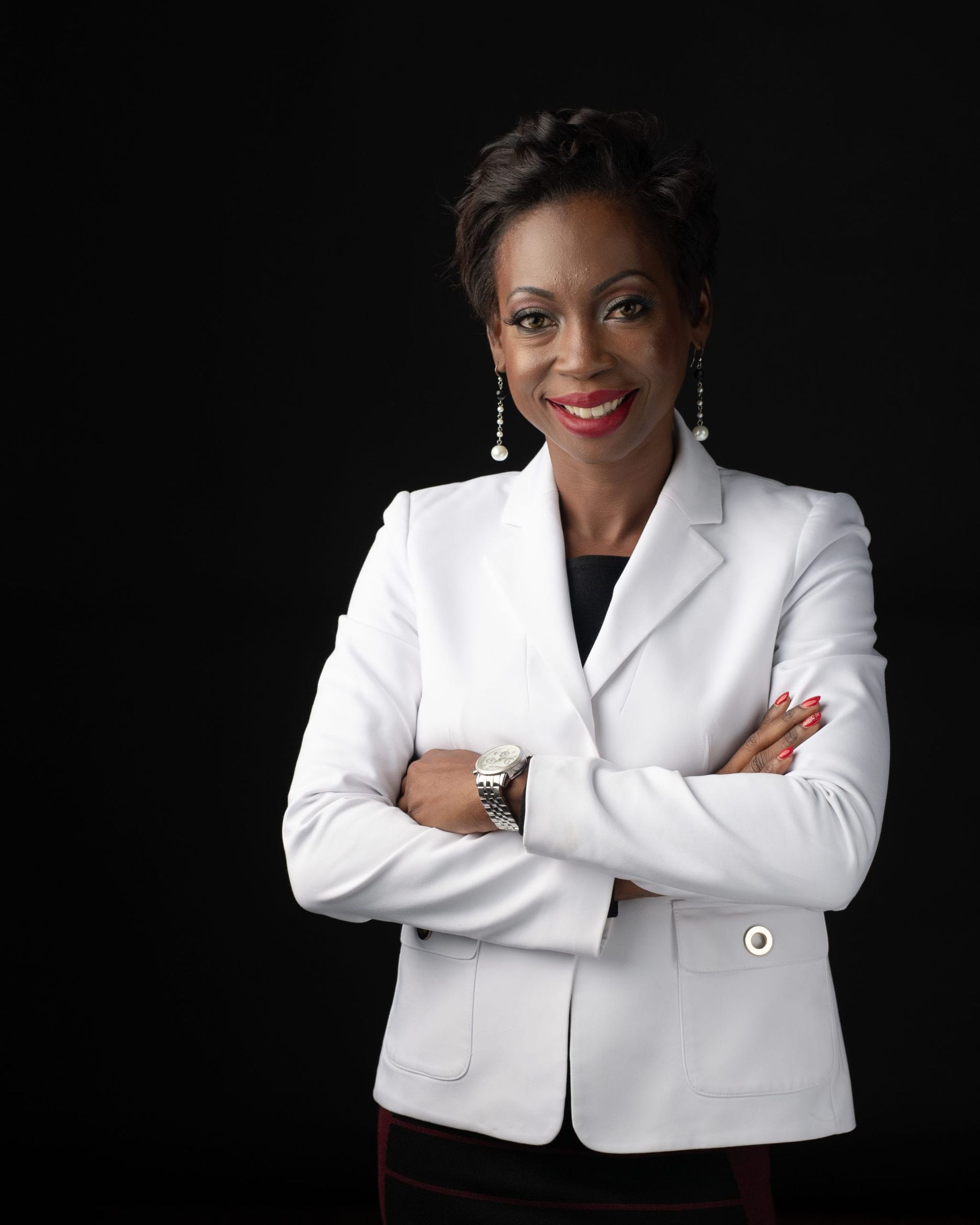 Woman in white blazer, arms crossed, smiles, against a black backdrop.