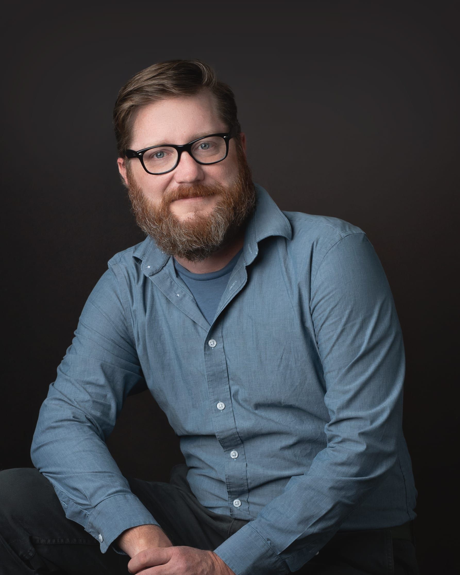 Man with beard and glasses, seated, smiling in front of a dark background, wearing a blue shirt.