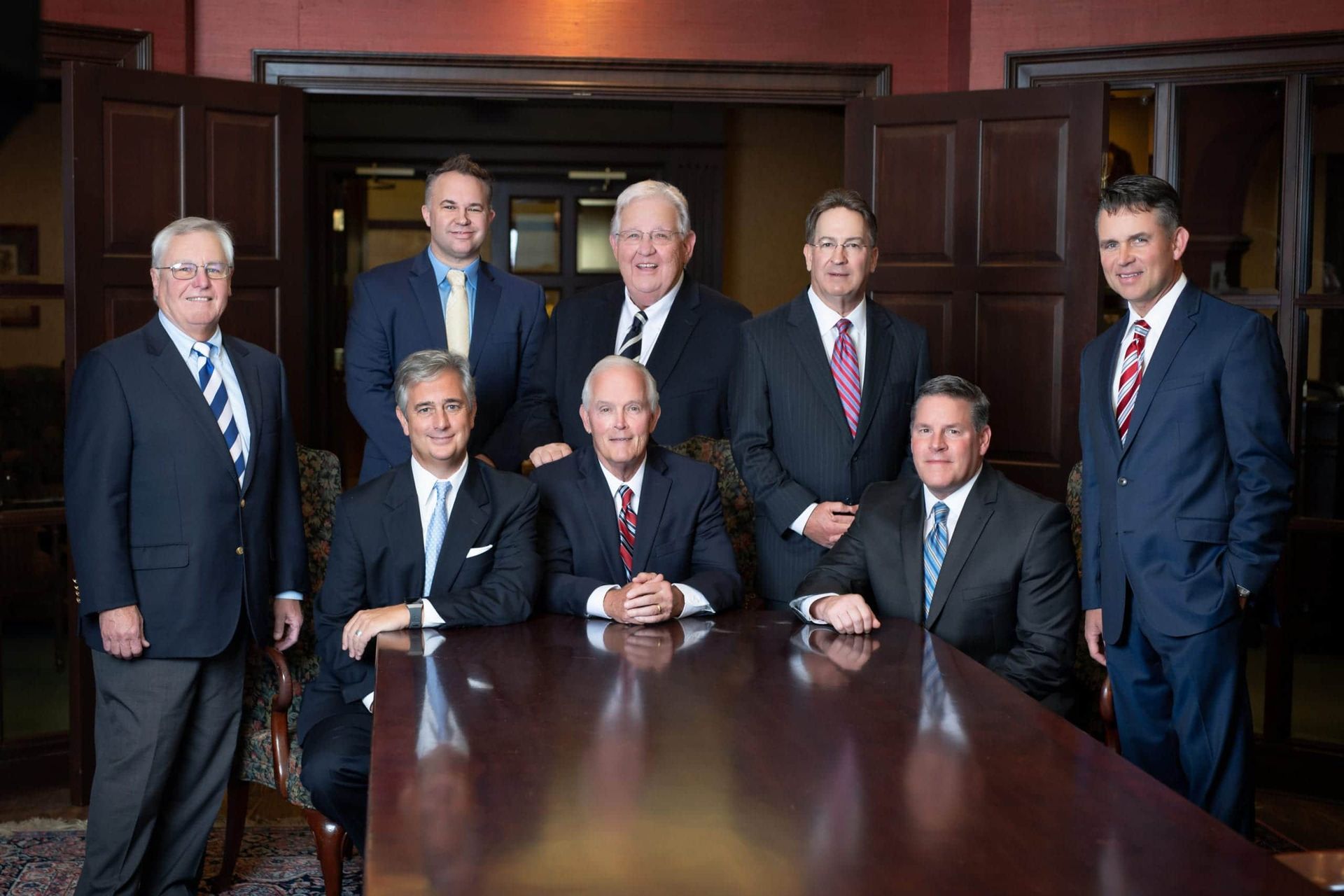 Group of eight men in suits, seated and standing around a large wooden table. Formal setting.