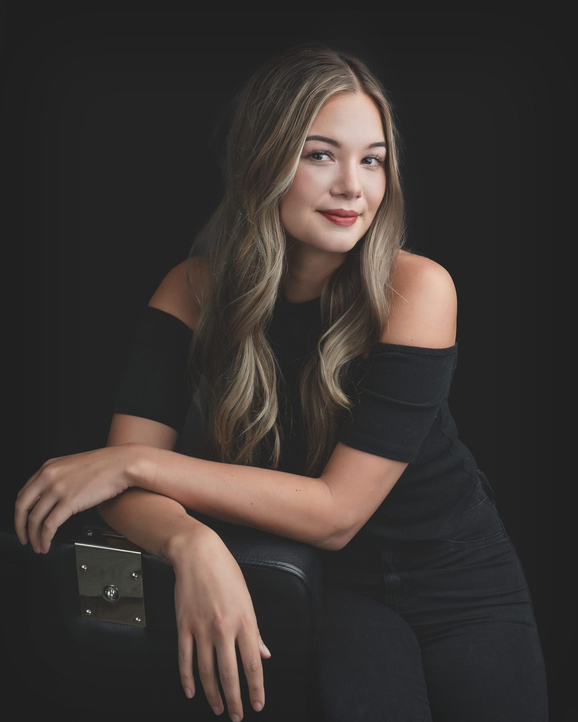 Young woman with long wavy blonde hair, wearing an off-the-shoulder black top, smiles, leaning on a dark object, black background.