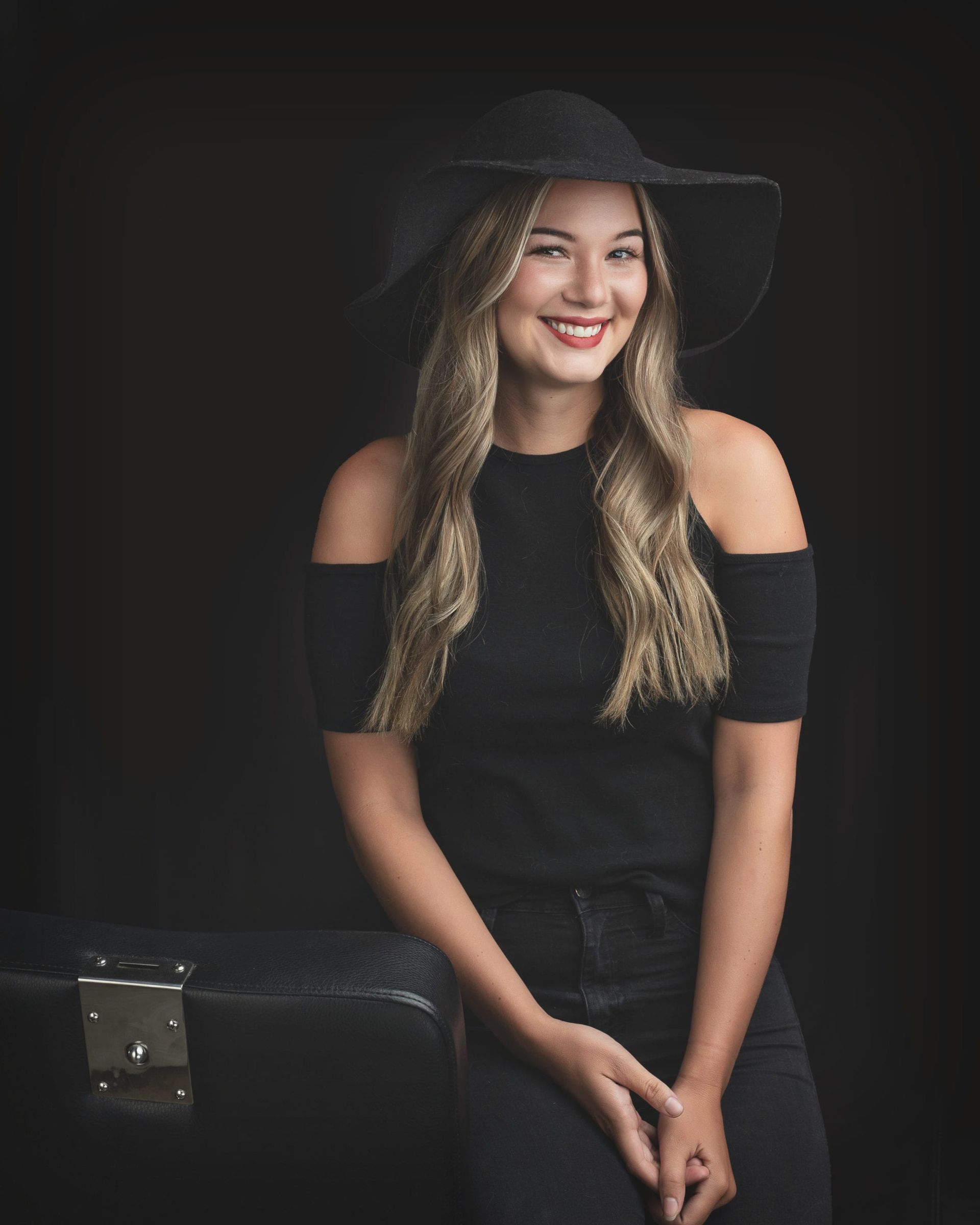 Woman in black hat and off-shoulder top smiles, leaning on a vintage suitcase against a dark background.