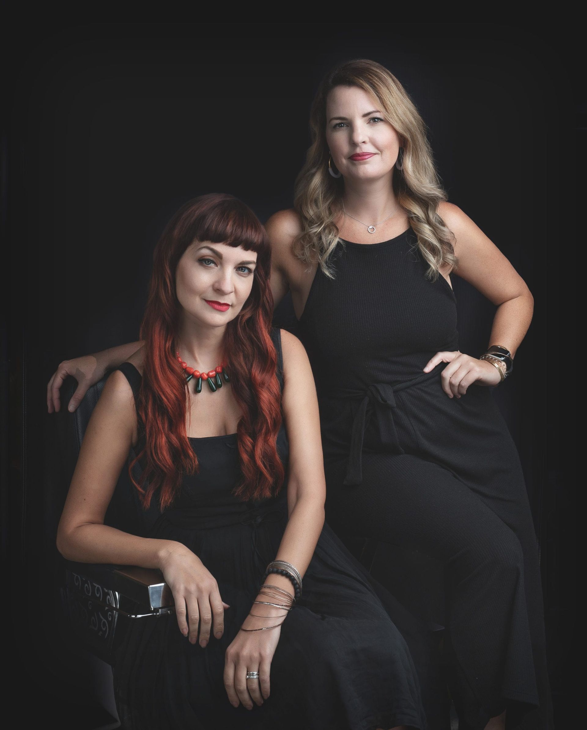 Two women in black dresses pose against a black backdrop; one sits, the other stands.