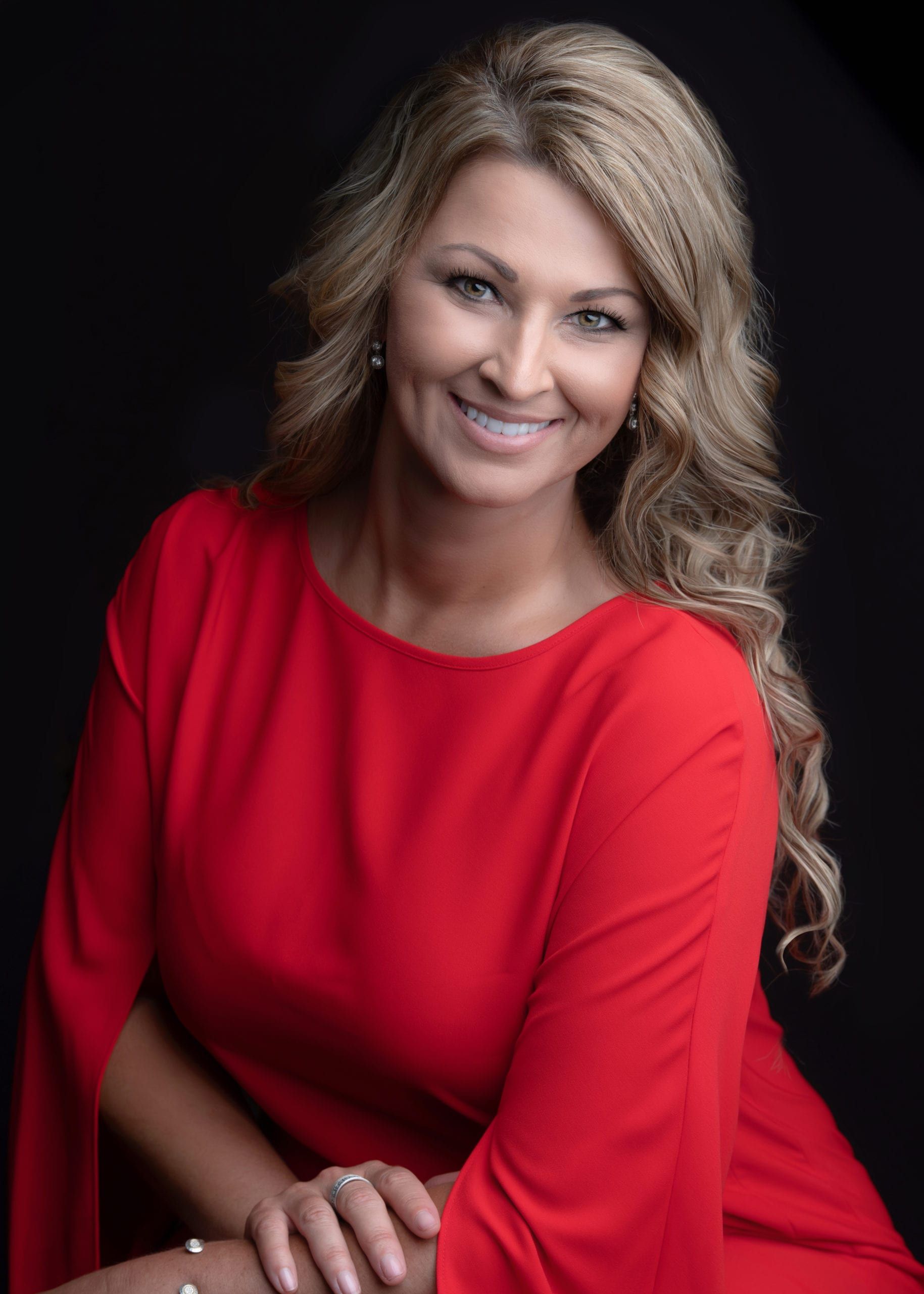 Woman in a red dress smiles, seated against a black backdrop; blonde hair, professional headshot.
