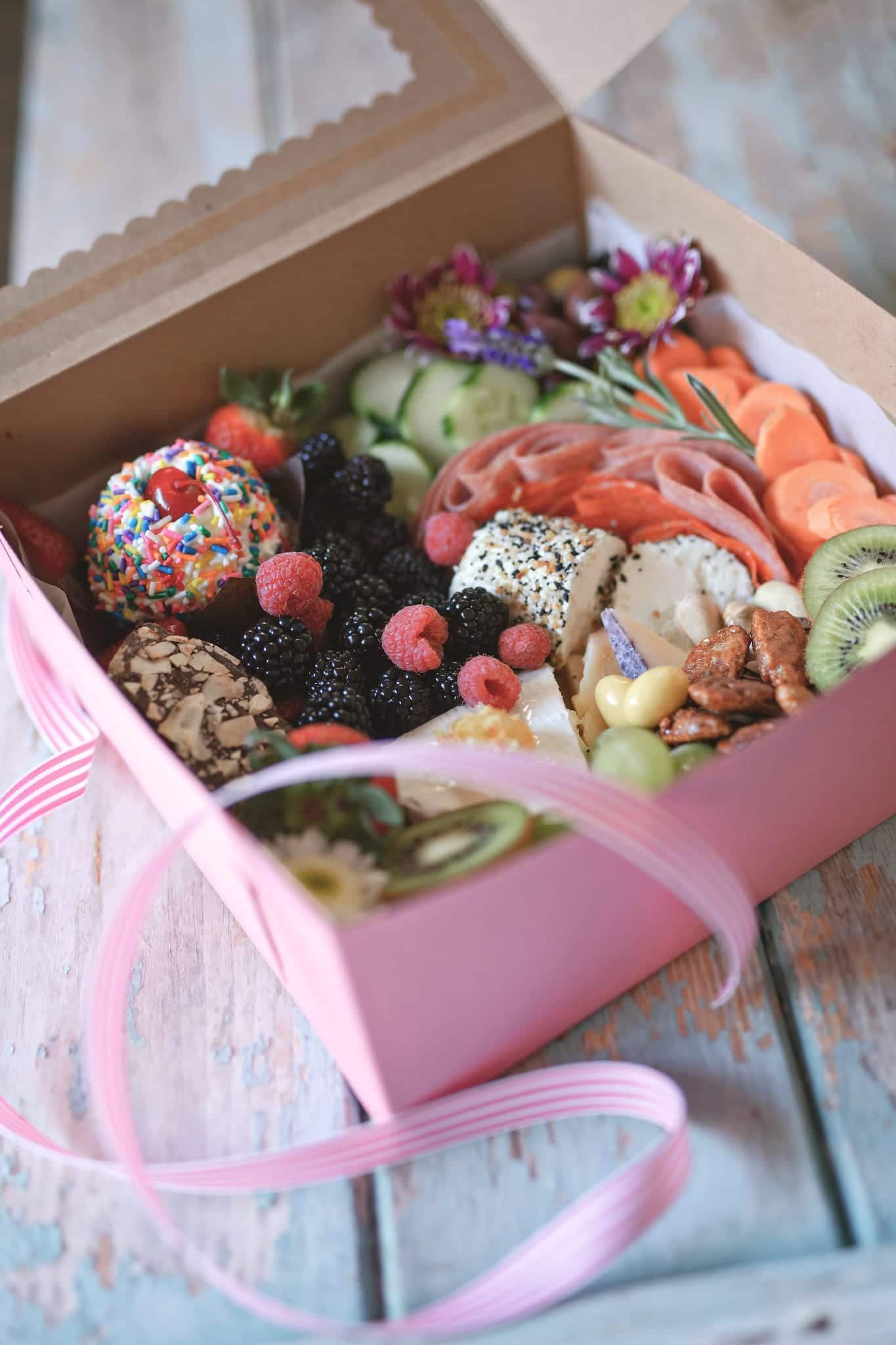 Charcuterie box with fruit, cheese, crackers, and flowers; pink ribbon.