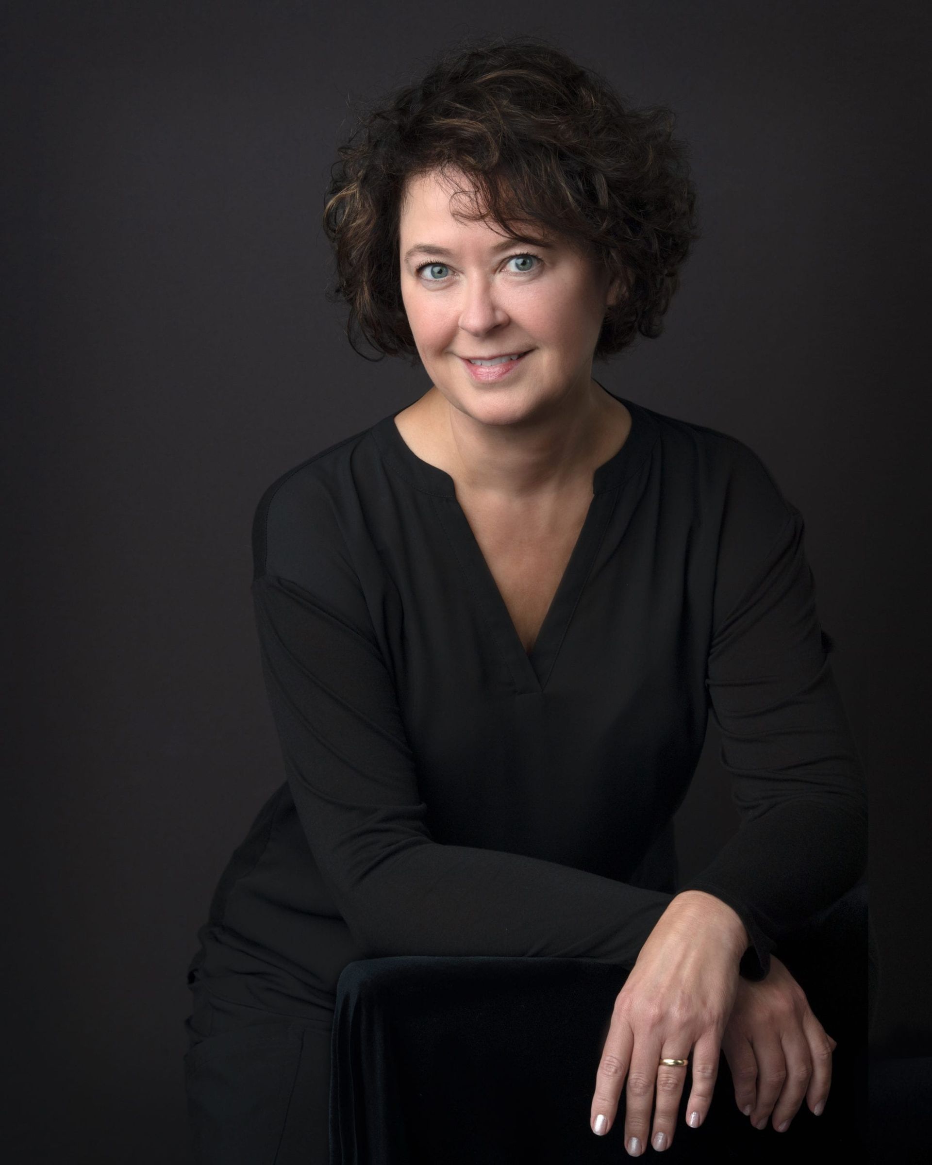Woman with curly brown hair, wearing a black shirt, smiles, leaning on a chair.