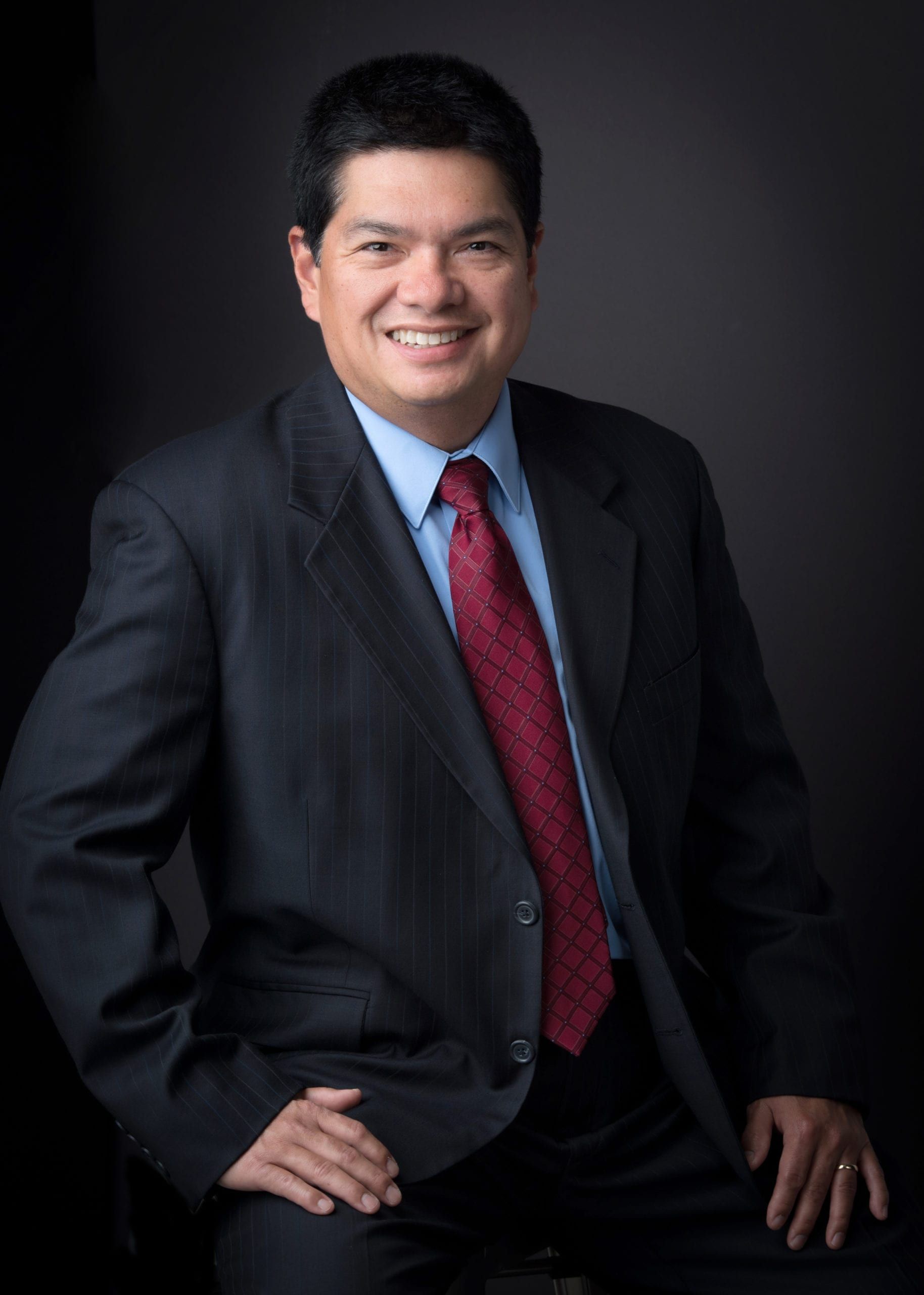 Man in suit, smiling, with a red tie, posed against a dark background.