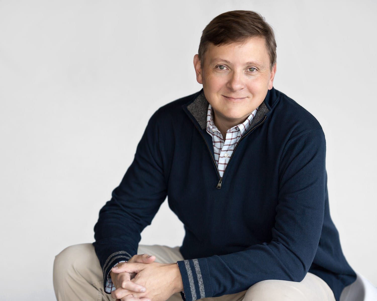 Man in navy sweater and khakis smiles, seated against a white backdrop.