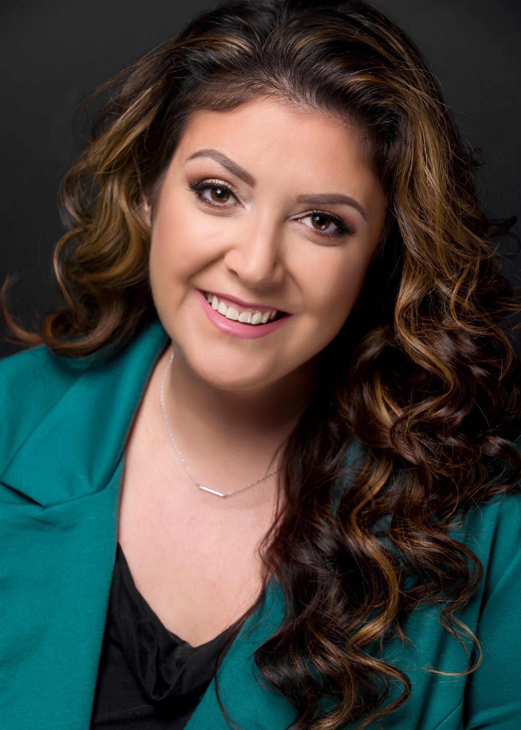 Woman with long, wavy brown hair smiles; wears teal blazer, black top, and silver necklace. Dark background.