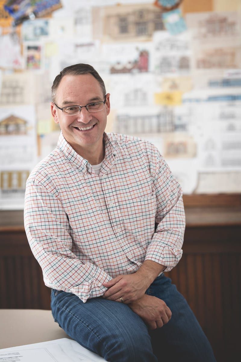 Man with glasses smiles, sitting on a ledge. He wears a plaid shirt and jeans, with architectural drawings behind him.