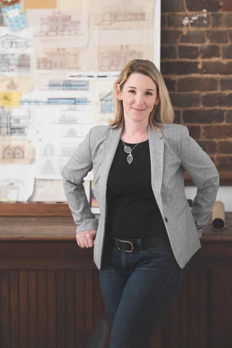 Woman in blazer, leaning on wooden counter. Blonde hair, jeans, and black shirt. Architectural drawings in background.