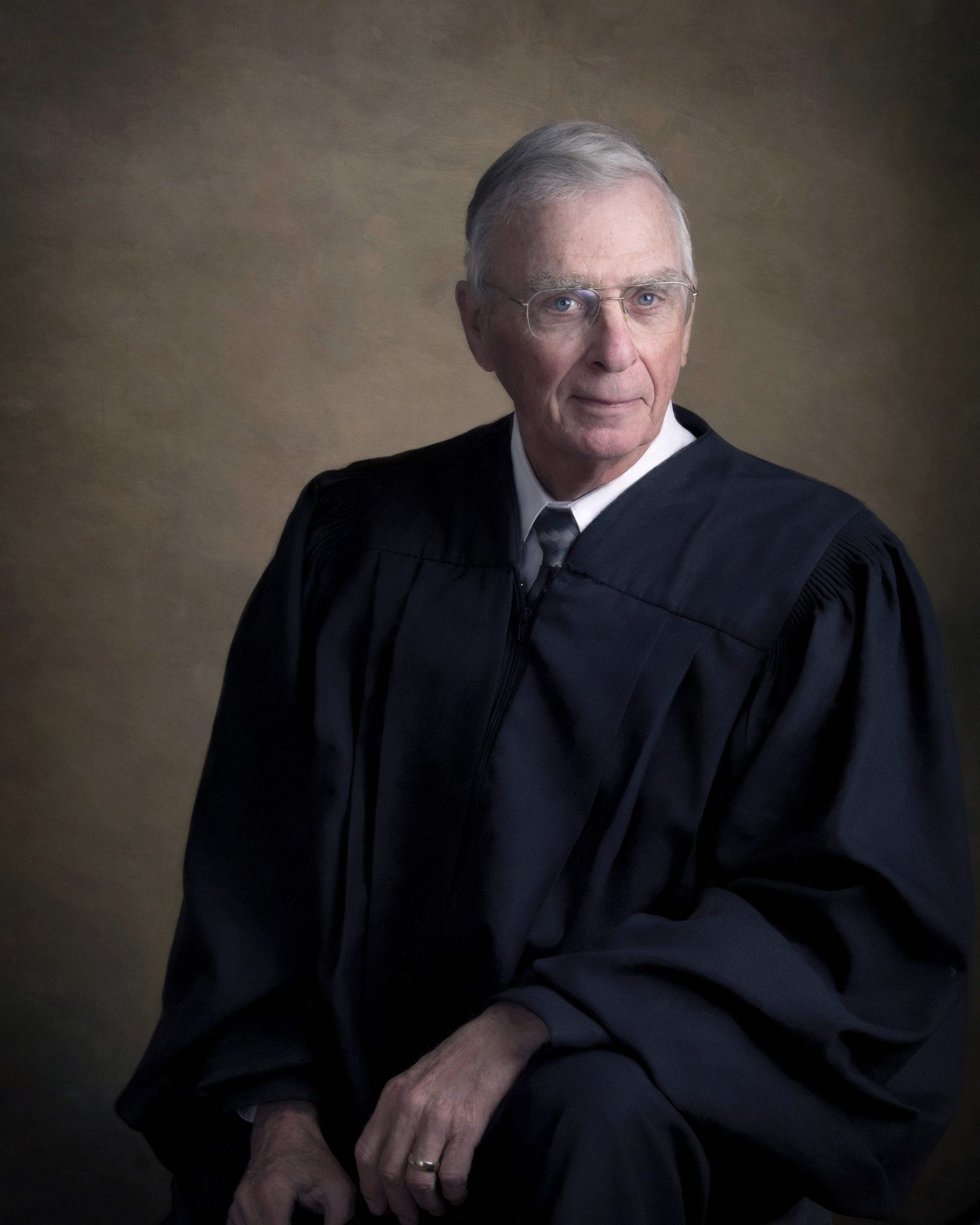 Judge in black robe, seated, looking to the right, against a brown backdrop.