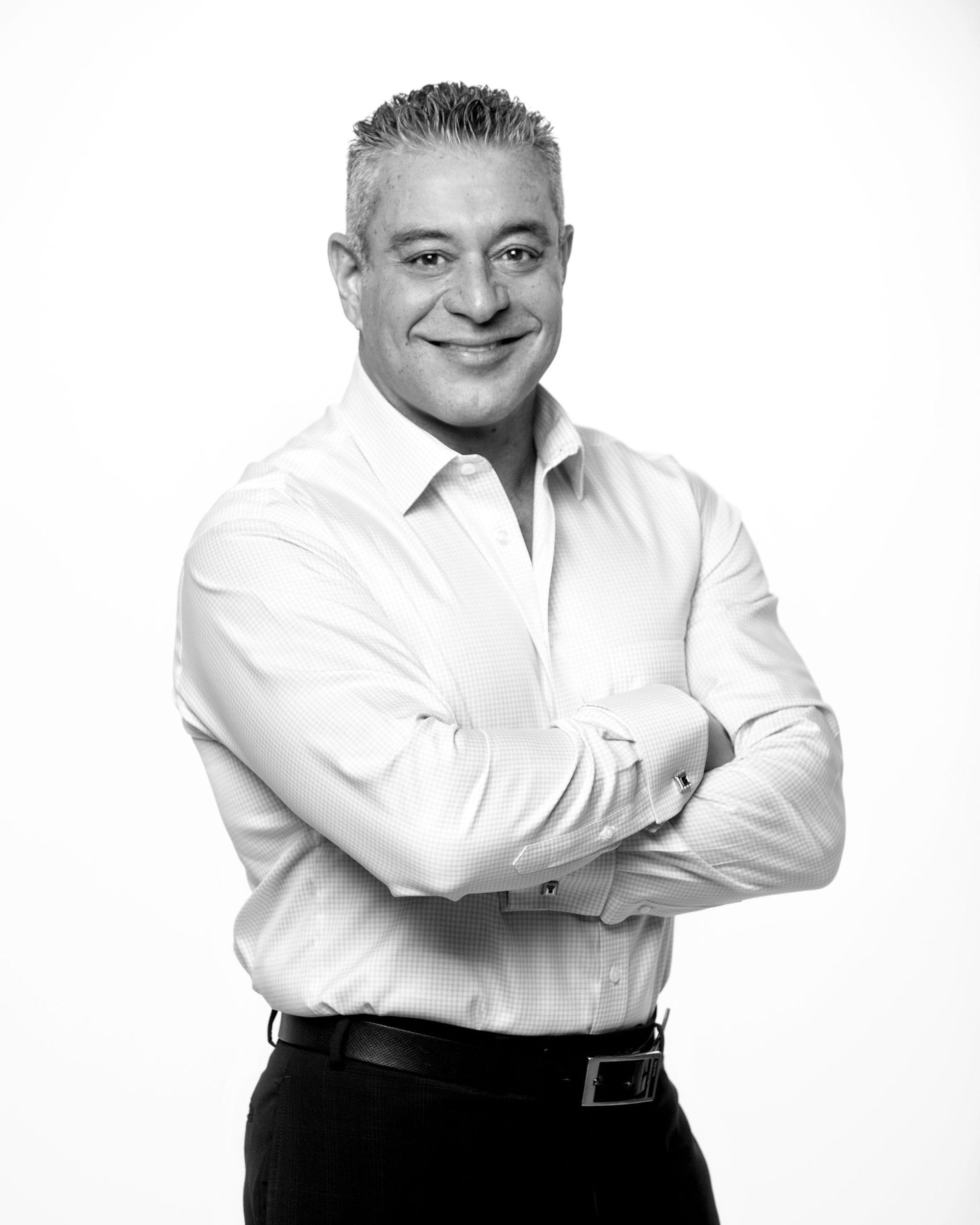 Man with graying hair, smiling, arms crossed, wearing a light-colored dress shirt and dark pants.