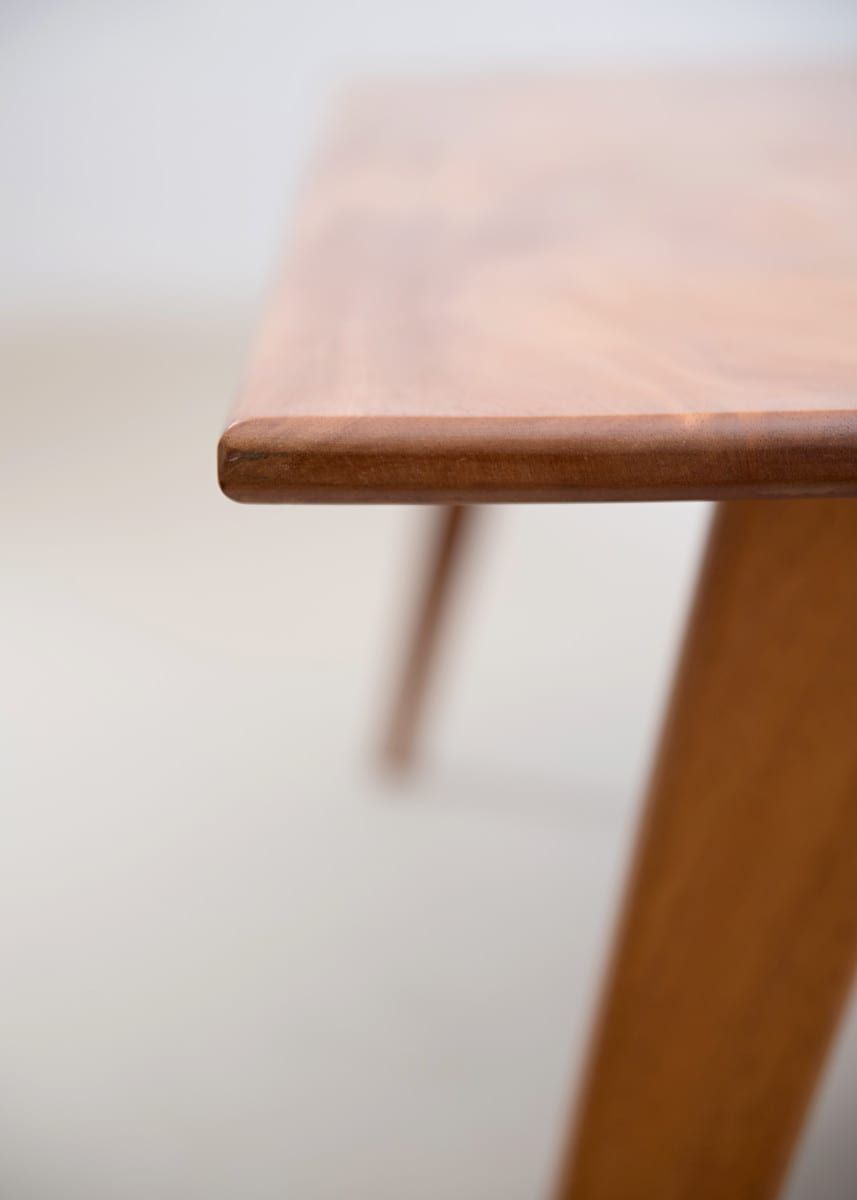 Close-up of a wooden table's corner, showing the tabletop edge and angled leg, against a white backdrop.
