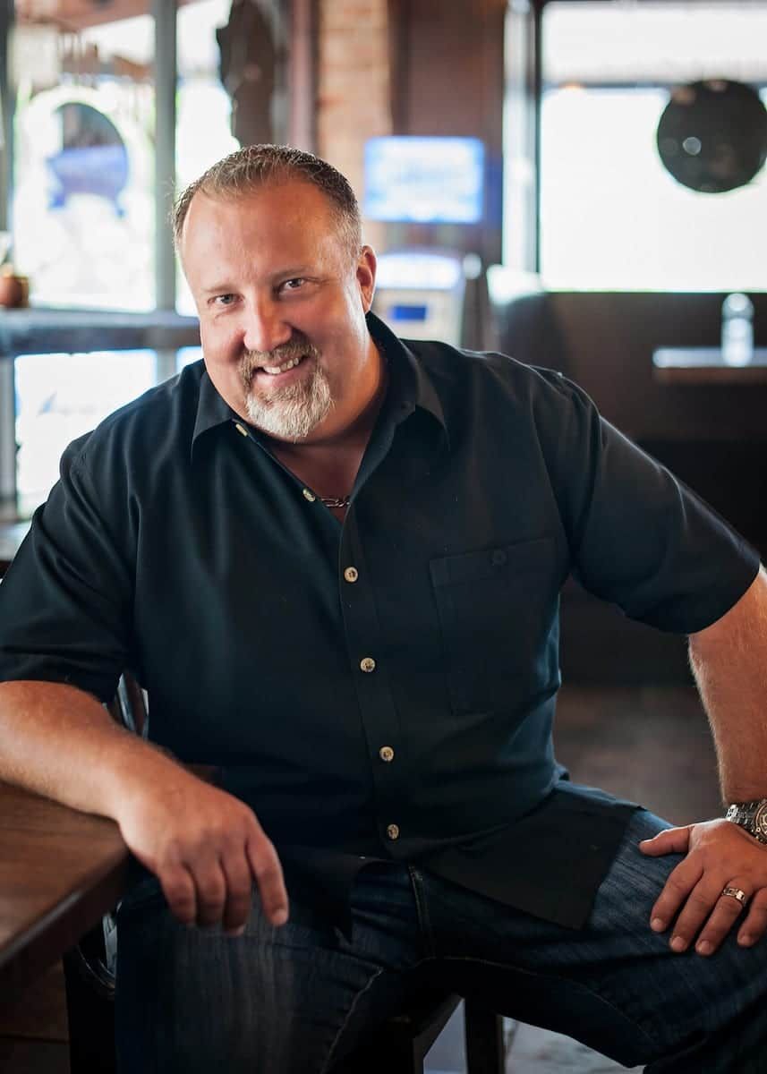 Man with beard smiles, seated at table in a restaurant, wearing black shirt, dark jeans.