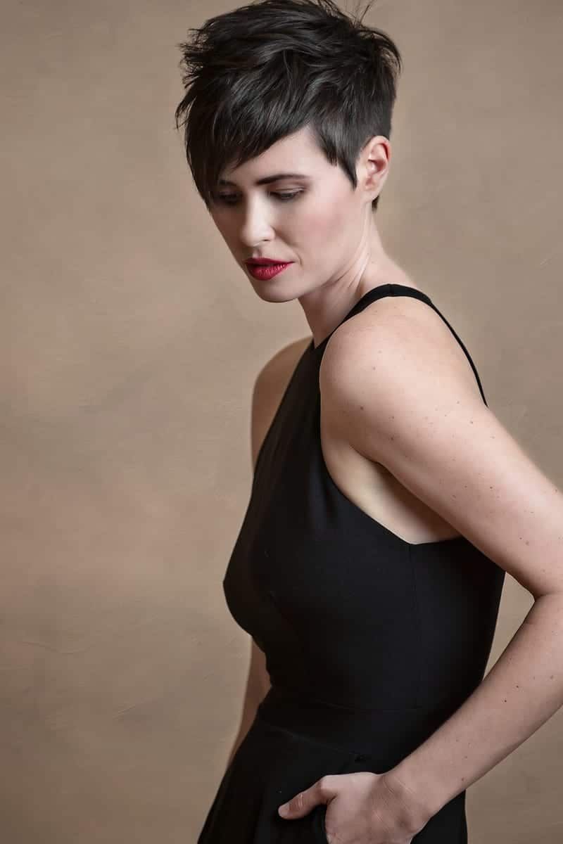 Woman with short dark hair wearing a black dress, looking down, hand in pocket. Neutral background.