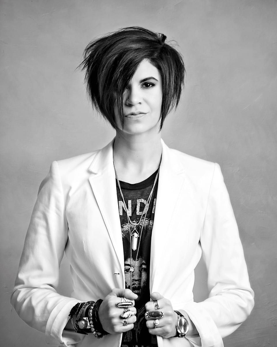 Woman in white blazer, short dark hair, wearing jewelry, neutral background.
