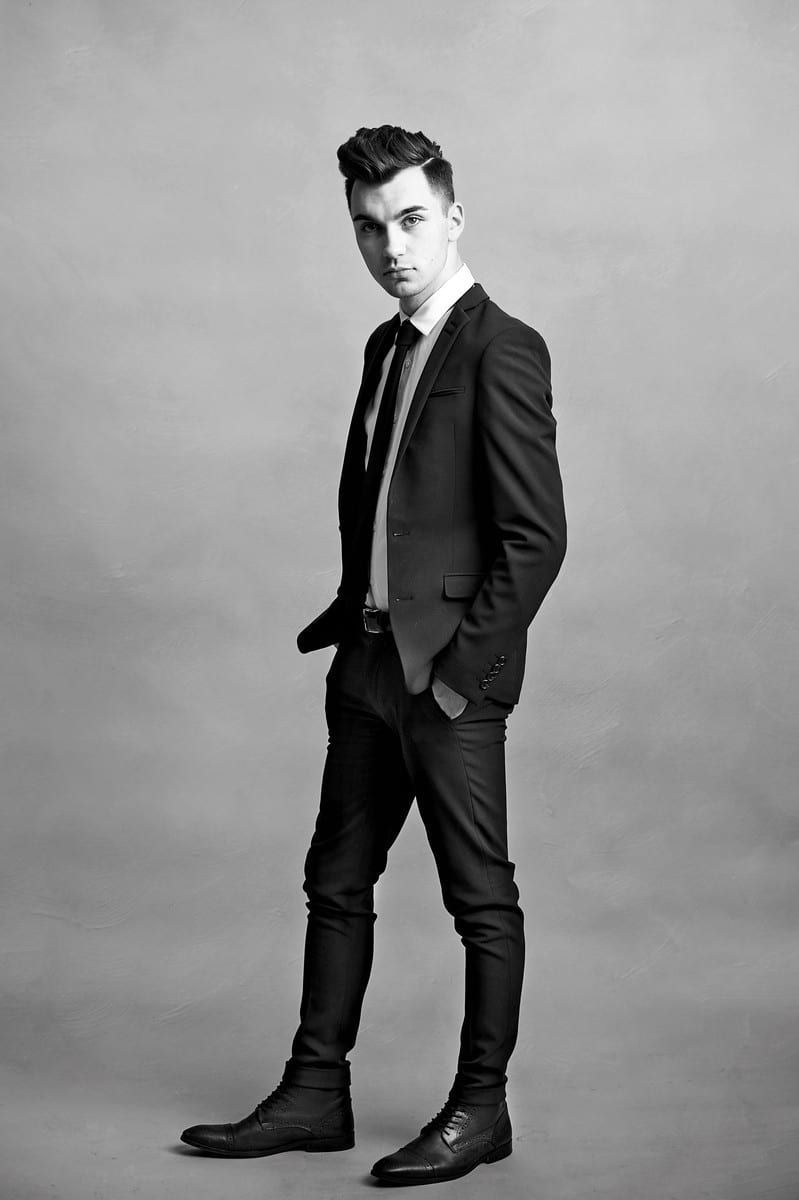 Young man in a black suit, tie, and boots, posing confidently with one hand in his pocket.