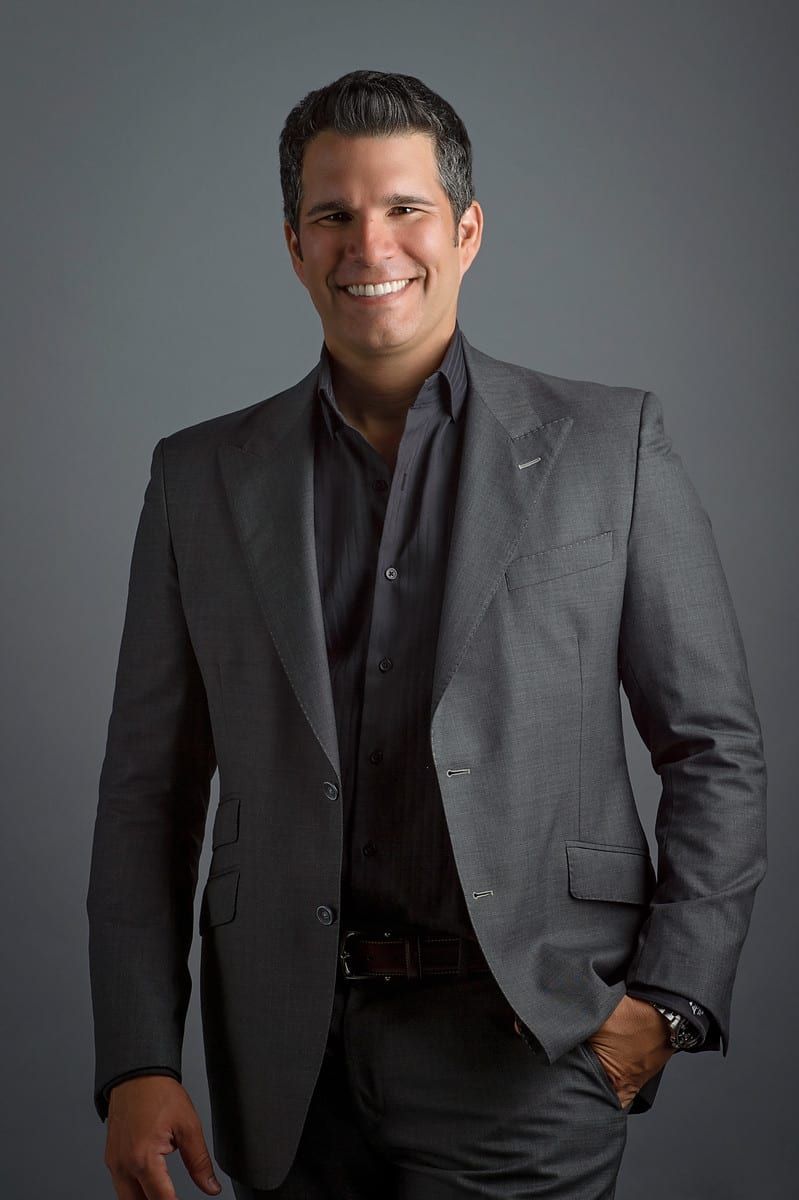 Man in gray suit smiling, hand in pocket, against a gray backdrop.